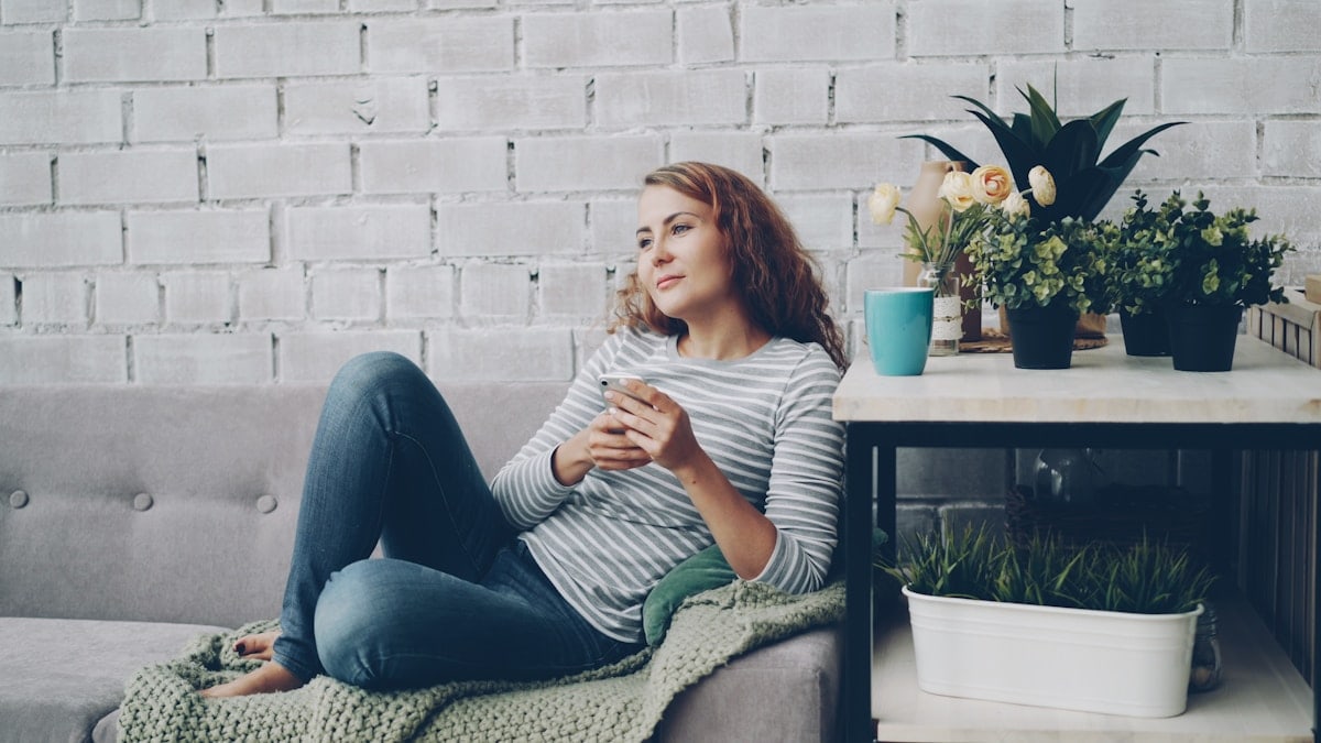 Attractive girl in casual clothing is using smartphone sitting on comfortable sofa in modern loft style apartment and smiling. Modern technology, interior and youth concept.